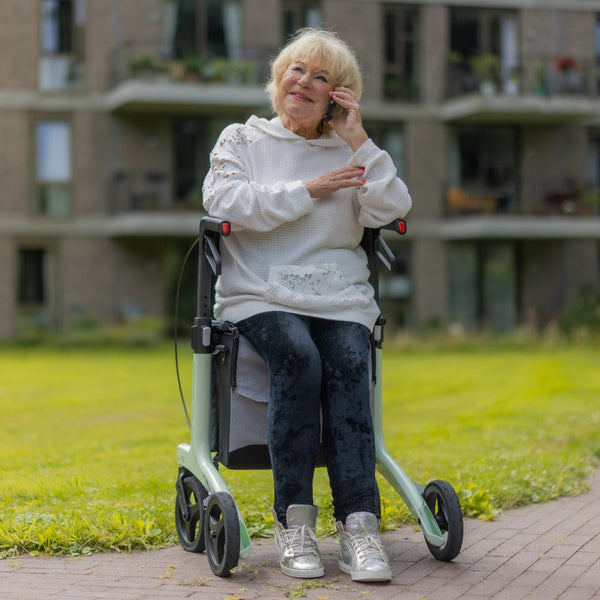 Elderly woman using a coal black carbon fibre Rollz Air rollator, talking on her mobile phone outside an apartment building, in use view.