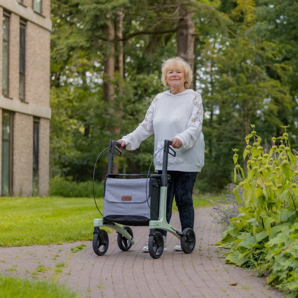 Older woman with light hair using a coal black carbon fibre rollator on a garden path with green plants, in use view.
