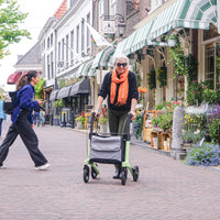 Older woman with white hair and orange scarf using a Rollz Air Carbon Fibre Rollator on a shop-lined street, in use view.