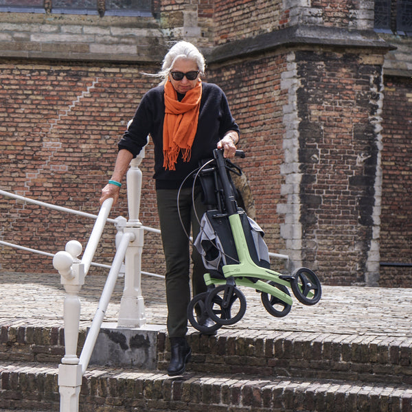 Rollz Air Carbon Fibre Rollator in Coal Black being carried by an older woman down stone steps outdoors, in use view.