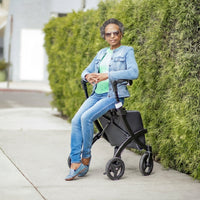 Woman sitting on a Rollz Flex 2-in-1 Rollator and Shopping Trolley on pavement, front view.