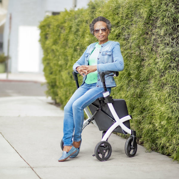 Rollz Flex 2-in-1 Rollator and Shopping Trolley in Jungle Green with woman sitting, wearing jeans, denim jacket and sunglasses, side view.