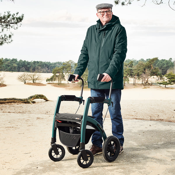 Older man in green jacket and cap outdoors with Rollz Motion Performance All-Terrain Rollator and Wheelchair, in use, side view.