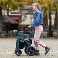 Woman in glasses and denim jacket using Rollz Motion Performance All-Terrain Rollator outdoors, trees and tables in background, side view.