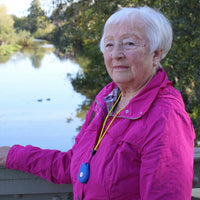 Elderly woman outdoors wearing Ravencourt Safer Walking GPS Locator by a riverside, trees in background, in use view.