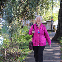 Older woman walking in park carrying brown bag with Ravencourt Safer Walking GPS Locator, in use.