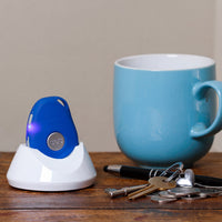 Ravencourt Safer Walking GPS Locator on charging dock with mug, keys, pen and coins on wooden surface, close-up.