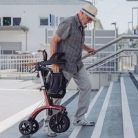 Man lifting Saljol Allround Bariatric Rollator with all-terrain tyres up outdoor steps, sunny day, in use.