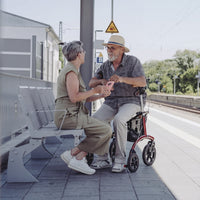 Older man seated on a Saljol Allround Bariatric Rollator at train station, speaking with woman holding smartphone, in use view.