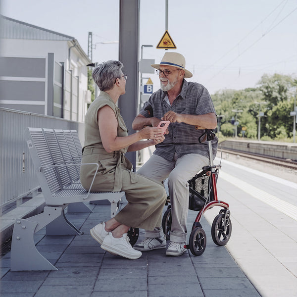Older man seated on a Saljol Allround Bariatric Rollator at train station, speaking with woman holding smartphone, in use view.