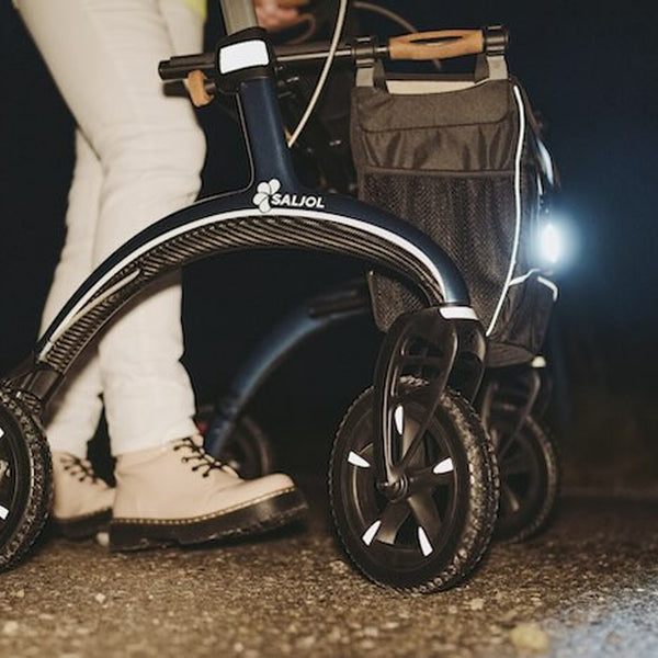 Person using a Saljol Bariatric Carbon Rollator with front light and black storage bag at night, in use view.