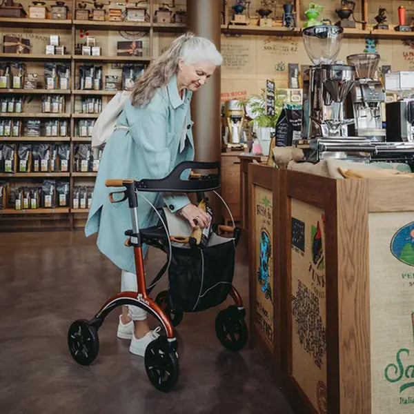 Older woman with long grey hair using a Saljol Carbon Rollator in a coffee shop, shelves visible behind her, in use view.