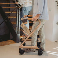 Person using a Saljol Indoor Rollator indoors while watching someone go up a wooden staircase, in use view.