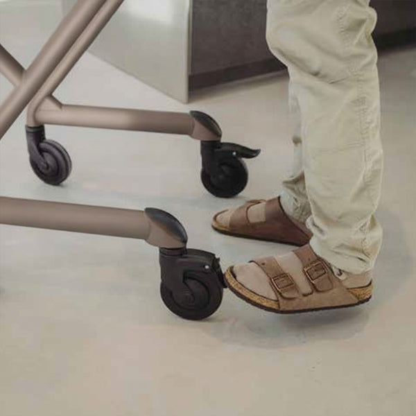 Person in beige sandals and trousers using the brake on a Saljol Indoor Rollator, in use view.