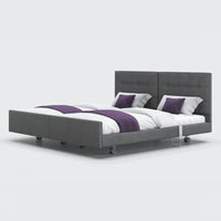 Dual profiling bed with divided headboard, white bedding, purple accents and wheels, front view.
