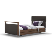 Upholstered profiling bed with wheels, white bedding, purple throw, and two pillows, side view.