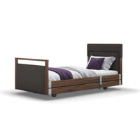 Opera Beds Signature Upholstered Profiling Bed with dark wood frame, white bedding, purple pillows and wheels, front view.
