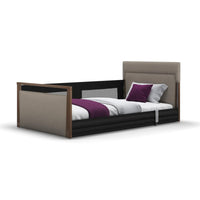 Solo SafeSide Upholstered Profiling Floor Bed with wood trim, black rails, white bedding and purple pillows, front view.