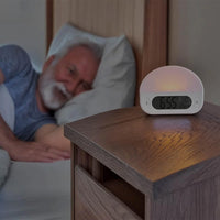 Lifemax Sunrise Alarm Clock displaying 6:55 on wooden nightstand beside elderly man in bed, in use view.