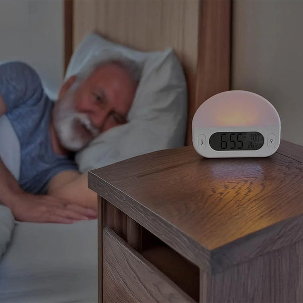 Lifemax Sunrise Alarm Clock displaying 6:55 on wooden nightstand beside elderly man in bed, in use view.