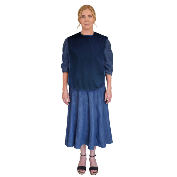 Woman wearing Care Designs tabard style adult bib over denim dress and black sandals, hair tied back, white background, front view.