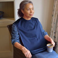 Woman wearing Care Designs Tabard Style Adult Bib sitting in chair holding a mug, looking to the side, in use side view.