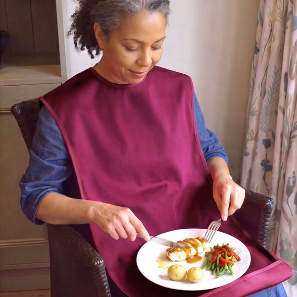 Woman wearing Care Designs Tabard Style Adult Bib eating a meal with fork and knife, seated at table, in use.