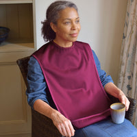 Woman wearing Care Designs Tabard Style Adult Bib, sitting in chair holding mug, looking out window, in use view.