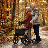 Elderly couple walking by a lake, man using Topro Olympos All Terrain Rollator, benches and trees along path, in use view.