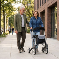 Older woman using a Topro Troja 5G Premium Rollator for support while walking outdoors with partner, in use.