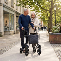 Older couple walking outdoors, one using Topro Troja 5G Premium Rollator for support, trees and shops in background, in use view.