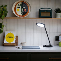 The Daylight Company TriSun Therapy Lamp on a modern desk with laptop, coffee cup, book and pen, shelves and white tile wall in background, front view.