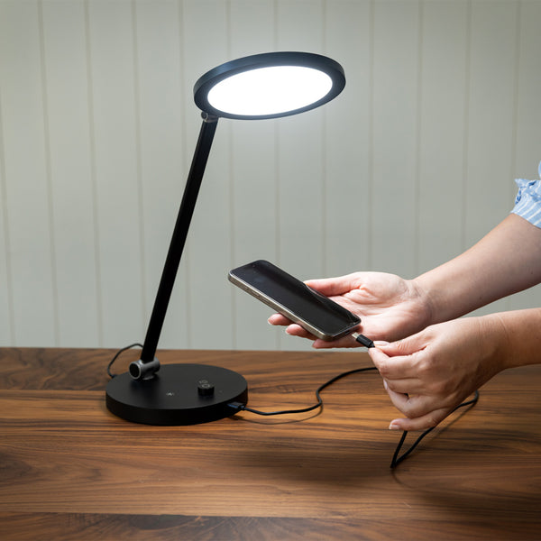 TriSun Therapy Lamp by The Daylight Company with phone charging via USB port on a wooden table, in use.