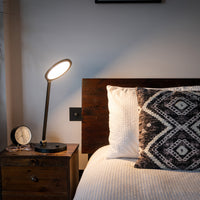TriSun Therapy Lamp with adjustable brightness on a nightstand beside a bed with white bedding and patterned pillow, in use view.