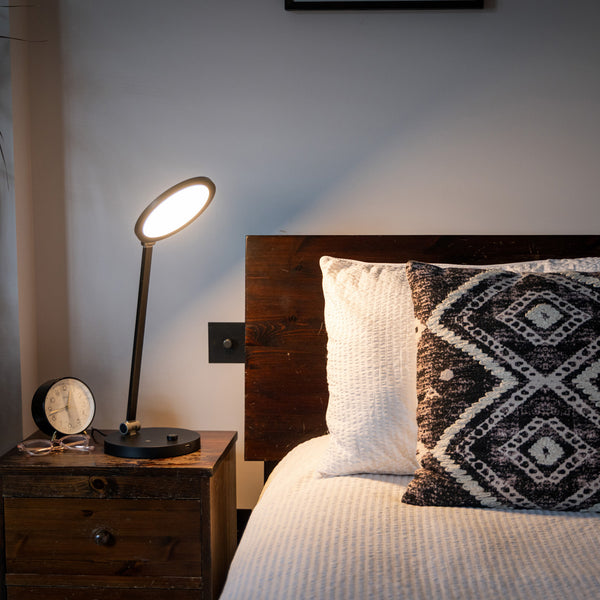 TriSun Therapy Lamp with adjustable brightness on a nightstand beside a bed with white bedding and patterned pillow, in use view.