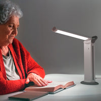 Elderly woman reading at a table under The Daylight Company Twist 2 Portable Lamp, in use view.