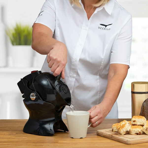 Pouring hot water from a Uccello Designs Uccello Kettle into a white mug on a wooden table with pastries and jar, in use view.