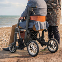 Able2 Uplivin Glide 2-in-1 Rollator & Wheelchair in use by the beach, one person seated and one standing, both wearing warm clothes, side view.